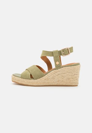 Oliven grøn ruskind kile sandal med flettet tov platform, åben tå, ankelrem og guldlås på hvid baggrund.