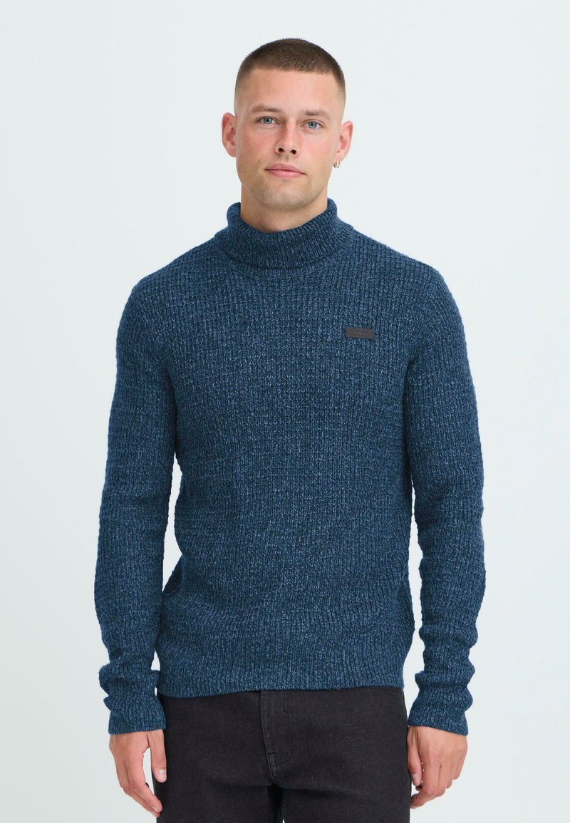 Temno moder pleteno pulover s turtleneck iz teksturirane tkanine in dolgimi rokavi, ki ima majhno črno etiketo na prsih.