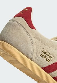Baskets Adidas Japan en daim beige avec languette rouge au talon, bandes rouges, logo doré et semelle extérieure en caoutchouc beige texturée.