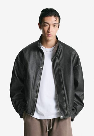 Black leather bomber jacket with a textured finish, silver hardware, and a snap closure, worn over a white t-shirt and beige sweatpants.