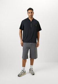 Sort kortærmet polo med halvlukning, brystlomme; parret med grå denimshorts. Modellen bærer grå sneakers med hvide accenter.