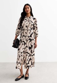 Midi-Kleid in einem beigen und schwarzen abstrakten Muster. Mit langen Ärmeln, durchgehender Knopfleiste, einem Gürtel in der Taille und Seitenschlitzen. Schwarze Handtasche und Schuhe.