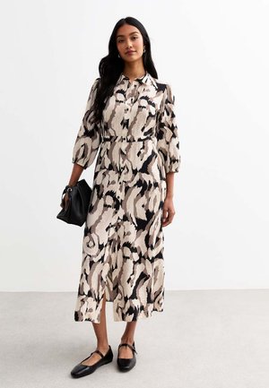 Abito midi con un motivo astratto beige e nero. Presenta maniche lunghe, chiusura con bottoni sul davanti, una cintura in vita e spacchi laterali. Borsa nera e scarpe.