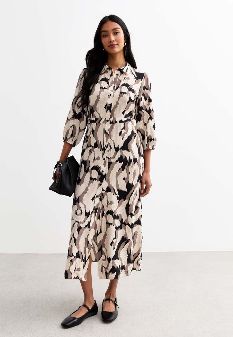 Midi-Kleid in einem beigen und schwarzen abstrakten Muster. Mit langen Ärmeln, durchgehender Knopfleiste, einem Gürtel in der Taille und Seitenschlitzen. Schwarze Handtasche und Schuhe.