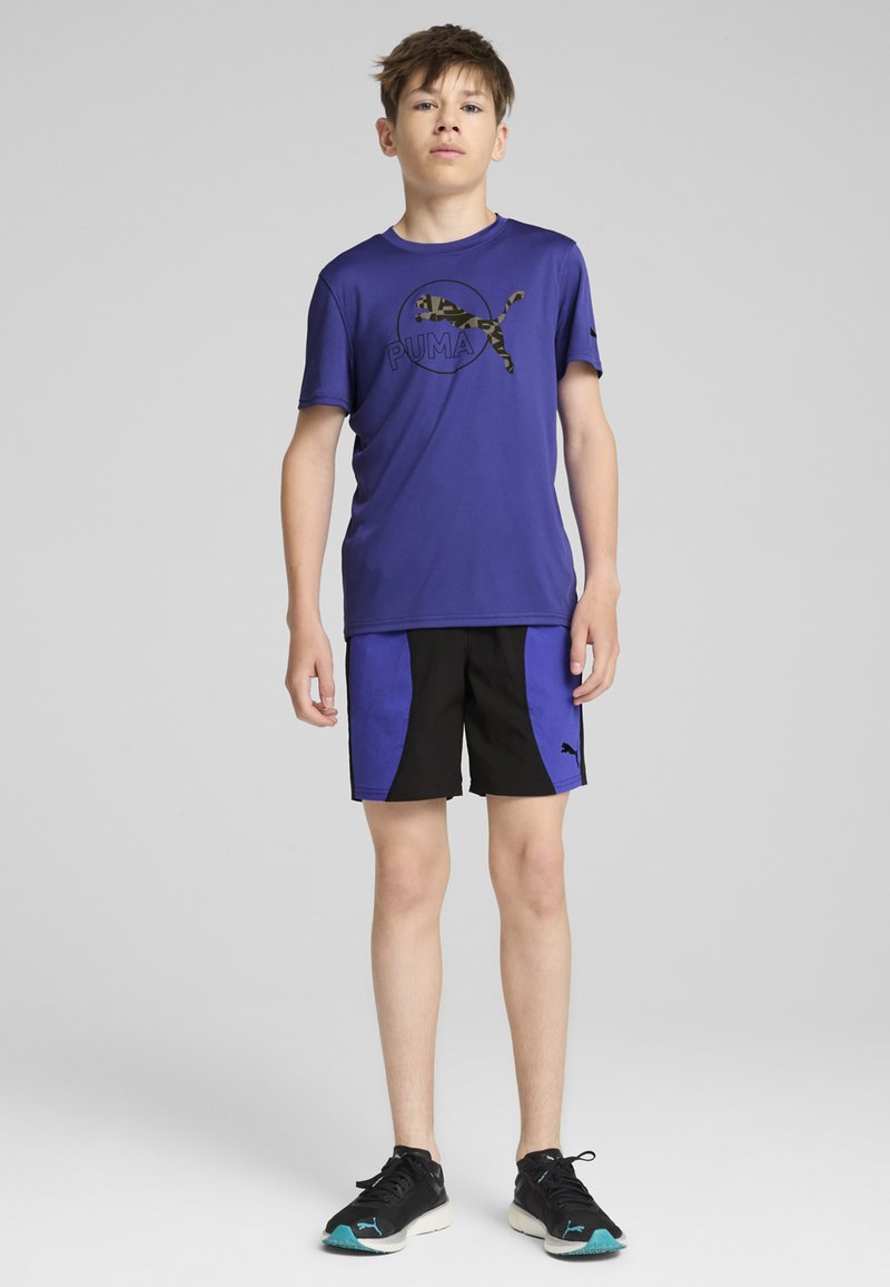 Puma lila sportt-shirt med svart logotyp, i kombination med svarta shorts med blå paneler. Bärs med svarta träningsskor.