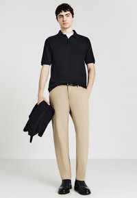 Polo nero, pantaloni beige, tiene un maglione nero. Le scarpe sono stivaletti neri. Sfondo semplice, modello in piedi.