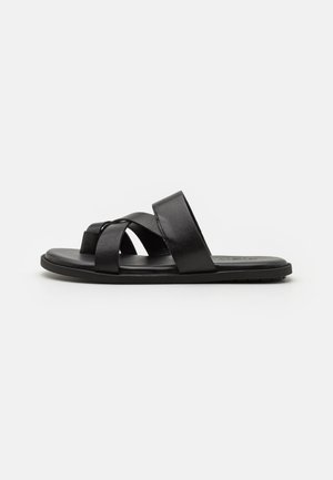 Sandal cu talpă plată din piele neagră, cu curele încrucișate și o textură netedă. Design simplu, cu detalii minimaliste.