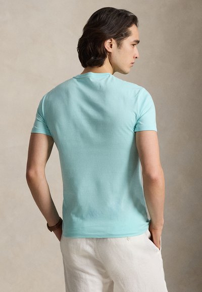 Άνδρας με σκούρα μαλλιά που φοράει ανοιχτό μπλε κοντομάνικη μπλούζα και λευκό παντελόνι, στέκεται με τα χέρια στις τσέπες κι έχει την πλάτη προς το μέρος μας.