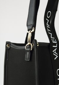 Fekete vászontáska bőr kiegészítőkkel, aranyszínű csattal és texturált pánttal, amelyen "VALENTINO" felirat található.