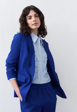 Γυναίκα με σγουρά σκούρα μαλλιά που φοράει μπλε σακάκι, ταιριαστό παντελόνι και ανοιχτό γαλάζιο ριγέ πουκάμισο με βολάν, ποζάροντας με τα χέρια στις τσέπες.