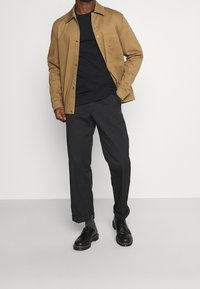 Giacca button-up beige sopra un maglione nero, abbinata a pantaloni scuri e scarpe nere. La giacca ha una tasca sul petto e una trama liscia.