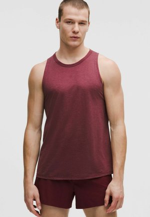 Junger Mann, der ein ärmelloses bordeauxrotes Tanktop und passende bordeauxrote Shorts trägt, vor einem schlichten hellen Hintergrund stehend.