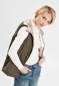 Gilet sans manches vert olive avec un intérieur doré brillant et fermeture éclair, porté par-dessus une chemise blanche à manches longues et un jean bleu.