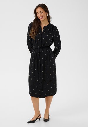 Femme en robe midi noire à manches longues boutonnée, avec un petit motif géométrique et une ceinture, portant des talons slingback noirs, souriante, les mains derrière.