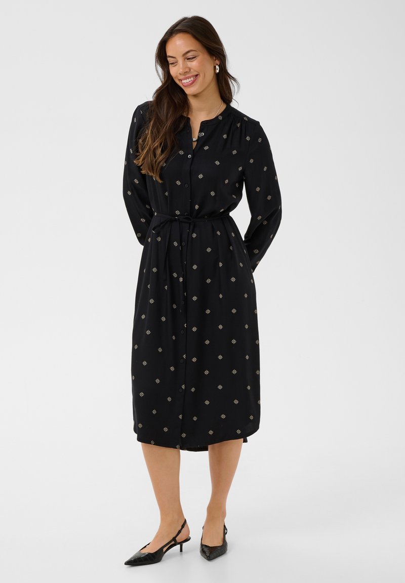Femme en robe midi noire à manches longues boutonnée, avec un petit motif géométrique et une ceinture, portant des talons slingback noirs, souriante, les mains derrière.