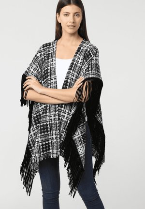Mujer con cabello largo y oscuro, vistiendo un chal de flecos a rayas blanco y negro sobre una blusa blanca y jeans azules, de pie con los brazos cruzados.