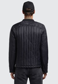 Black padded jacket with vertical quilted design, lightweight material, and snap closure detail. Worn with dark jeans and a black cap.