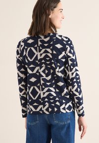 Kobieta w granatowo-beżowej, geometrycznej wzorzystej bluzce z długim rękawem, w towarzystwie niebieskich dżinsów, stojąca na tle jasnego, jednolitego tła.