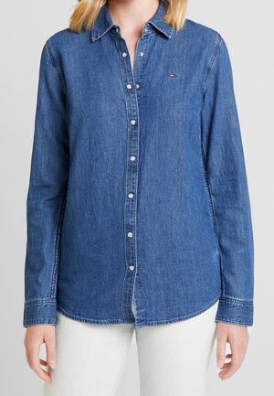 Frau trägt ein blaues, langärmeliges Jeanshemd mit Knöpfen und kleinem Logo auf der Brust sowie weiße Hose, steht vor einfarbigem Hintergrund.