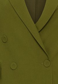 Olivgrön dubbelknäppt jacka med en texturerad yta, som har två runda knappar och en slagkrage.