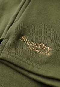 Tela verde oliva con textura acanalada que presenta un logotipo bordado en dorado de "SuperDry Athletics Co." en la parte inferior.