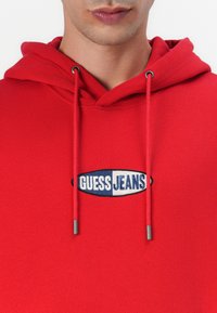 Red hoodie with drawstrings, featuring a blue and white embroidered "GUESS JEANS" logo on the chest. Fabric texture appears smooth.