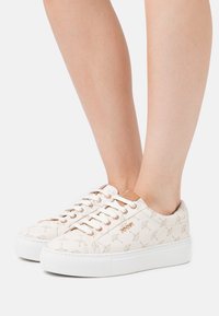 Kengät, joissa on teksturoitu kermaisen värinen päällinen, jossa on hillitty kuvio, valkoinen alustapohja ja pyöreät nauhat. Ruusukultaiset yksityiskohdat.