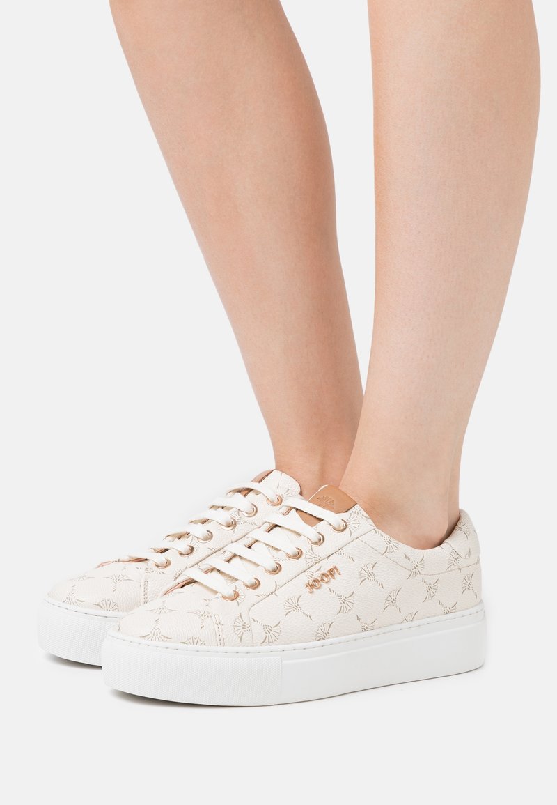 Kengät, joissa on teksturoitu kermaisen värinen päällinen, jossa on hillitty kuvio, valkoinen alustapohja ja pyöreät nauhat. Ruusukultaiset yksityiskohdat.