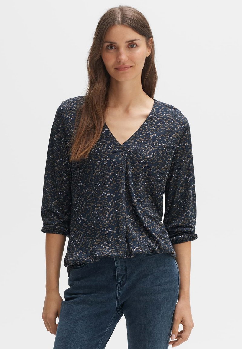 Bluse im Marine-Stil mit V-Ausschnitt, langen Ärmeln und lockerem Schnitt. Der Stoff hat eine glatte Textur und dezente metallische Akzente.