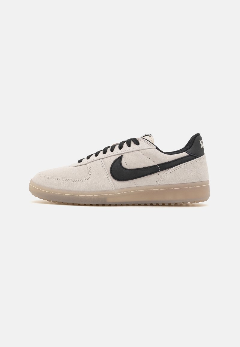 Baskets basses en daim beige avec le logo Nike noir, lacets noirs, languette de talon noire et semelle en gomme translucide sur fond blanc.