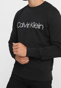 Černá mikina s bílým logem "CalvinKlein" přes hruď, kulatým výstřihem, dlouhými rukávy a žebrovanými manžetami a lemem.