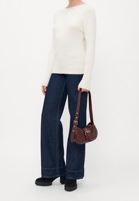 Personne portant un pull blanc à manches longues, un jean large sombre, des chaussures à plateforme noires, tenant un petit sac à bandoulière marron.