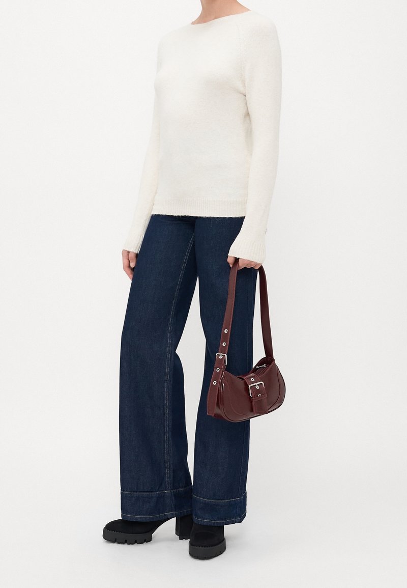 Personne portant un pull blanc à manches longues, un jean large sombre, des chaussures à plateforme noires, tenant un petit sac à bandoulière marron.