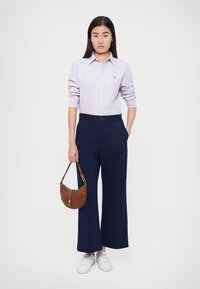 Gaiši violeta krekla jaka ar garām piedurknēm, tumši zili plašas bikses, baltas sporta kurpes un brūna somiņa ar rāvējslēdzēja detaļu.