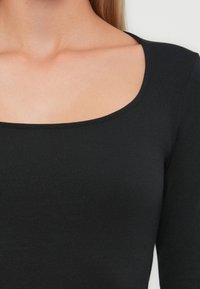 Haut noir à manches longues en tissu lisse et extensible, avec un col rond large et une coupe ajustée.