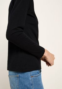 Person in einem schwarzen Langarmshirt und blauen Jeans, von der Seite gezeigt mit sanft gefalteten Händen vor sich.