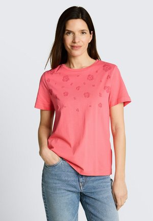 Kobieta z długimi brązowymi włosami, ubrana w różowy, krótki rękaw z subtelnymi kwiatowymi detalami i niebieskie dżinsy, stojąca z jedną ręką w kieszeni.