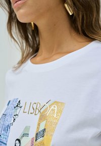 T-shirt en coton blanc avec un imprimé graphique coloré d'une scène de rue à Lisbonne, avec des lettres dorées et un motif de lanterne.