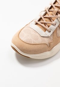 Närbild på beige och bruna sneakers som visar texturerad mockatå, mönstrat tyg, beiga skosnören och vit gummisula mot vitt bakgrund.