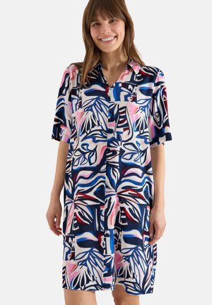 Femme souriante portant une robe à manches courtes boutonnée avec des motifs abstraits de feuilles et de vagues bleues, roses et blanches sur fond blanc.