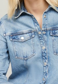 Light blue denim shirt featuring a button-down front, two chest pockets with flaps, and metallic accents on buttons and pockets.