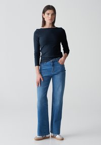 Schwarzes geripptes Langarmoberteil mit hohem Bootsausschnitt. Hellblaue Schlagjeans im klassischen Fünf-Taschen-Design. Legere weiße und marineblaue Sneakers.