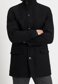 Černý vlněný kabát s vysokým límcem, zapínáním na knoflíky vpředu, dvěma předními kapsami a strukturovaným povrchem. Má rovný střih.