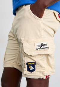 Pantaloni scurți cargo bej, fabricați dintr-un material ușor. Caracteristicile includ buzunare laterale, o etichetă cu logo-ul "Alpha Industries" și un emblemă "Airborne".