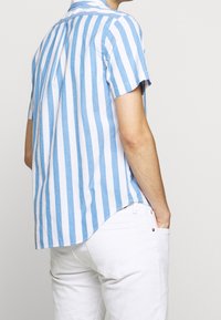 Košile s krátkým rukávem v světle modrých a bílých pruzích, vyrobená z lnu, s zakřiveným lemem a límečkem zapínaným na knoflíky. Nošena s bílými kalhotami.