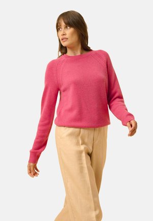 Maglione lavorato a maglia fucsia con maniche lunghe e polsini a coste, caratterizzato da un collo a giro e cuciture raglan. Abbinato a pantaloni beige.