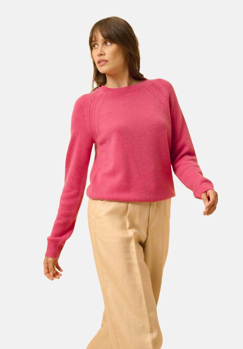 Maglione lavorato a maglia fucsia con maniche lunghe e polsini a coste, caratterizzato da un collo a giro e cuciture raglan. Abbinato a pantaloni beige.