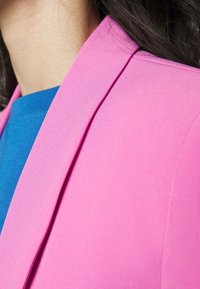 Jasnoróżowa marynarka o gładkiej fakturze, z klapami w kształcie litery V, noszona na niebieskiej bluzce. Projekt podkreśla czyste linie i minimalne detale.