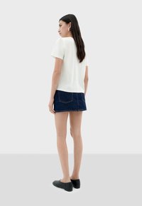 White short-sleeve top, paired with a dark denim skirt. Model wears black flats, seen from the back. Simple design, clean lines.