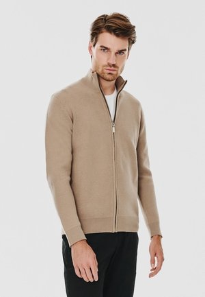 Beżowy sweter zapinany na zamek wykonany z miękkiego dzianinowego materiału, z wysokim kołnierzem, prążkowanymi mankietami i gładką fakturą, w połączeniu z ciemnymi spodniami.